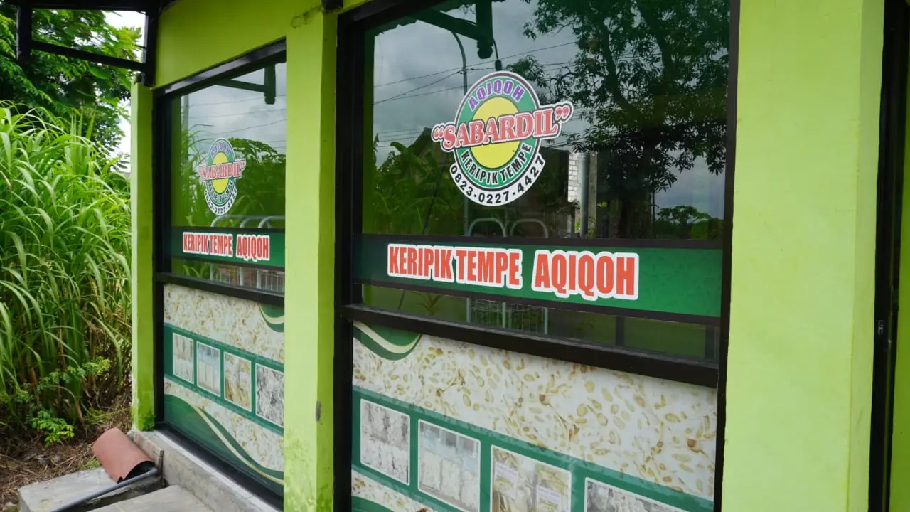 Tampak depan rumah produksi Keripik Tempe Aqiqoh, Ngampel, Kapas, Bojonegoro. Stiker logo UMKM tertempel di dinding kaca dengan tembok hijau.