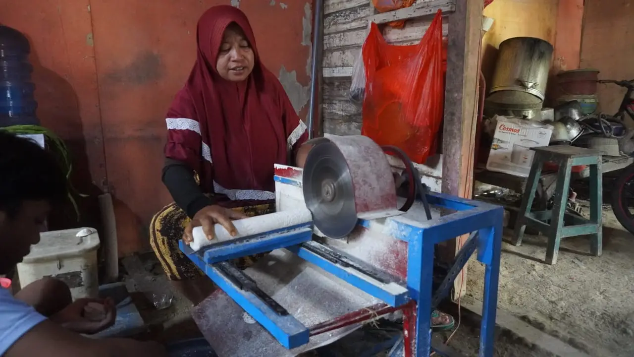Seorang pekerja duduk di balik alat pemotong tempe, tempe didorong dengan tangan ke pisau berputar untuk dipotong.
