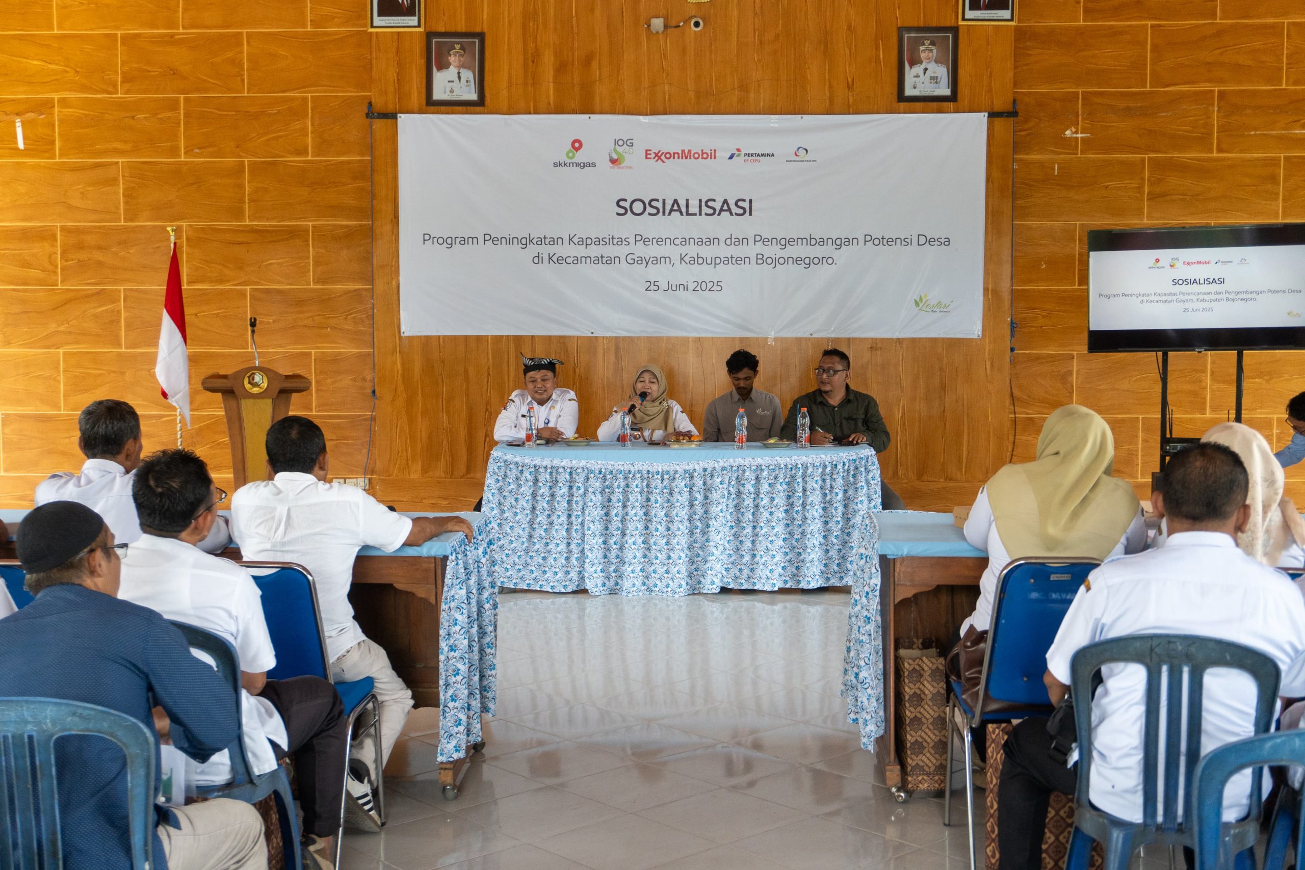 Tampak depan tedapat latar belakang banner putih berlogo ExxonMobil dan Pertamina di bagian atas tengah, serta logo Lestari Muda Indonesia di pojok kanan bawah, terpasang di tembok bermotif kayu coklat. Banner bertuliskan Sosialisasi Program Peningkatan Kapasitas Perencanaan dan Pengembangan Potensi Desa Kecamatan Gayam. Empat orang pembicara duduk di depan audience. Dua dari kiri, wanita memegang mikrofon dan berbicara.