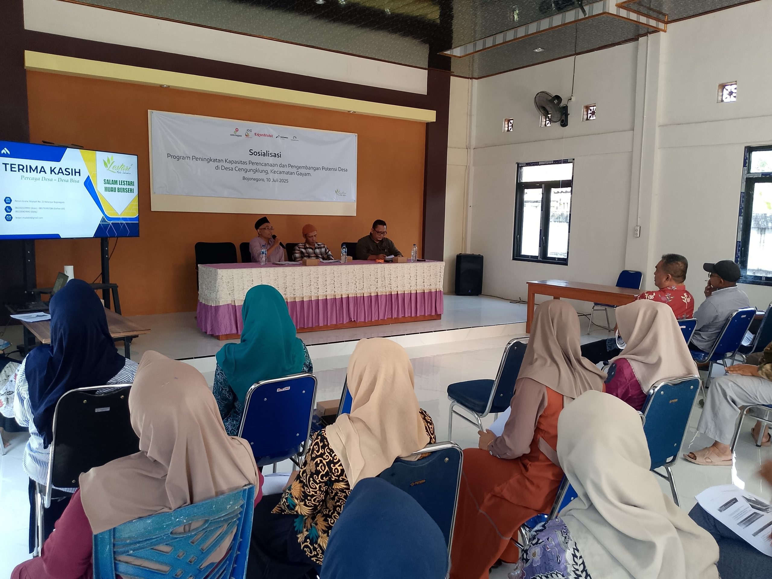 Pembicara duduk di kursi depan dibelakang meja panjang menghadap ke audiens. Beberapa audiens terlihat memperhatikan. Sebuah layar proyektor di samping meja pembicara menunjukkan materi. Di tembok belakang pembicara terdapat banner dengan tulisan "Sosialisasi Program Peningkatan Kapasitan Perencanaan dan Pengembangan Potensi Desa di Cengungklung, Kecamatan Gayam. Bojonegoro 10 Juli 2025."